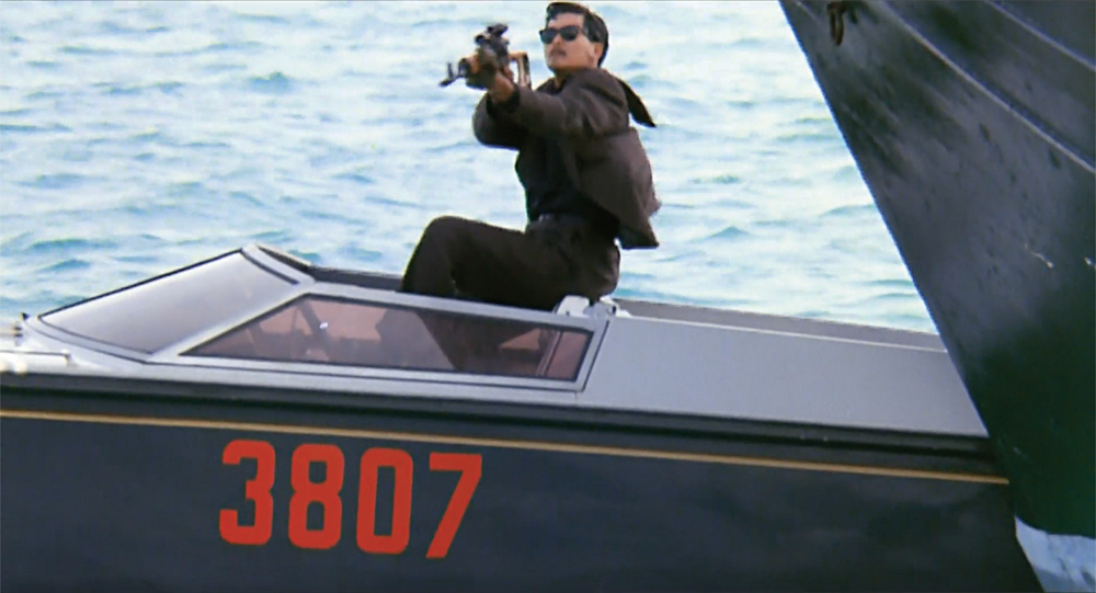 A man sits on the captain’s seat in a boat while aiming a gun into the foreground in The Killer.