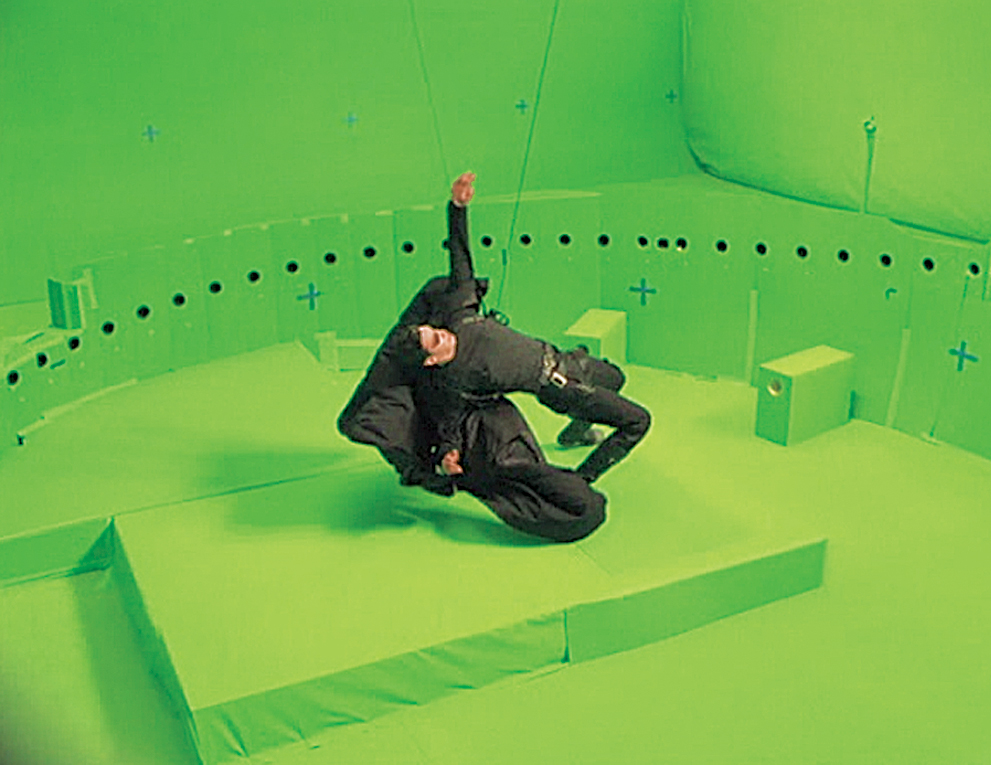 A man in a trench coat bending backwards at the knees is supported by wires on a circular set with many cameras surrounding the actor from The Matrix.