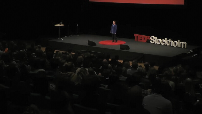 A screenshot of Greta Thunberg’s Ted X talk. She stands on a stage in an auditorium and a crowd is seated facing her.