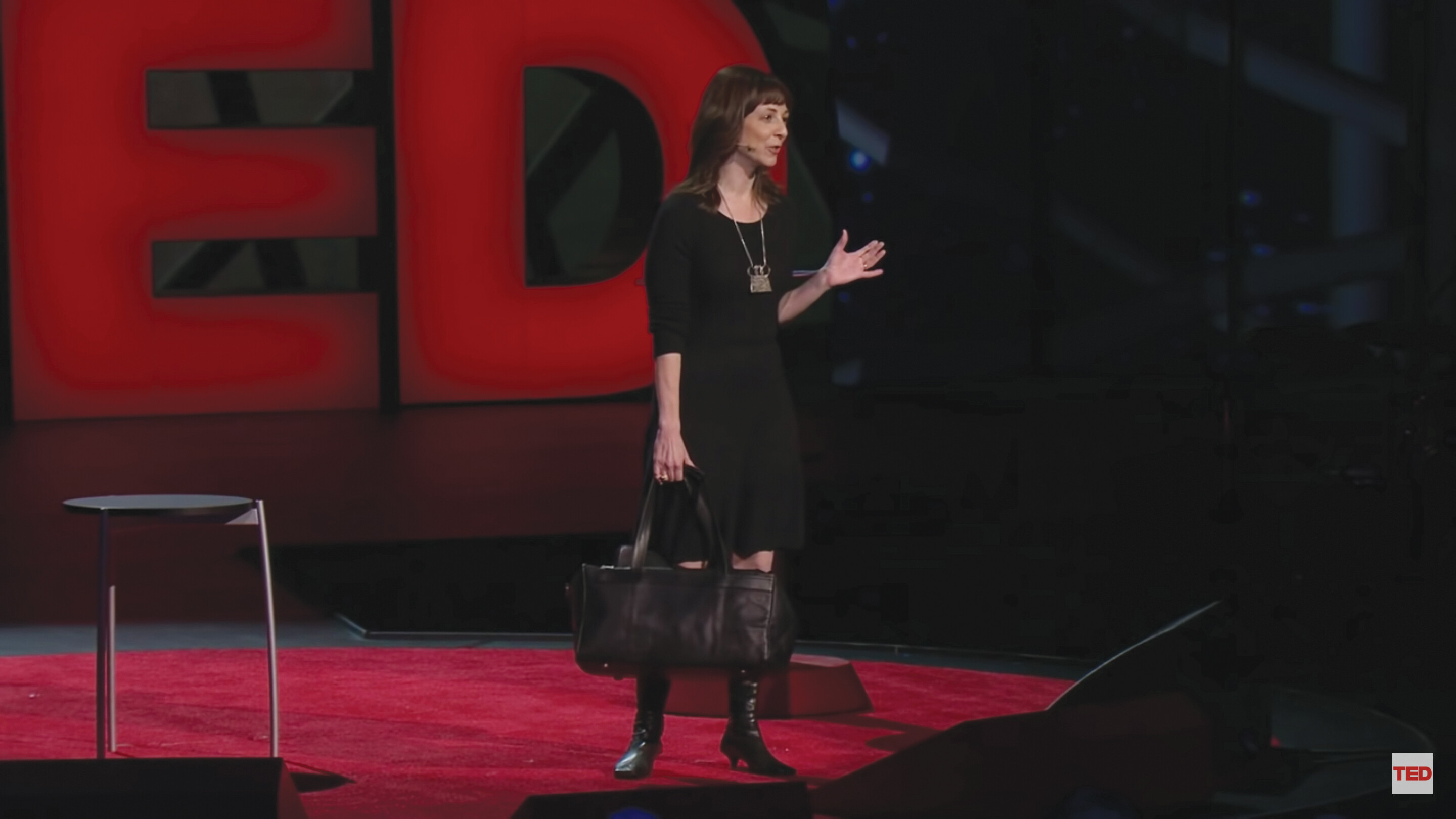 A screenshot of Susan Cain’s TED talk. She stands on a stage in an auditorium holding a black suitcase.