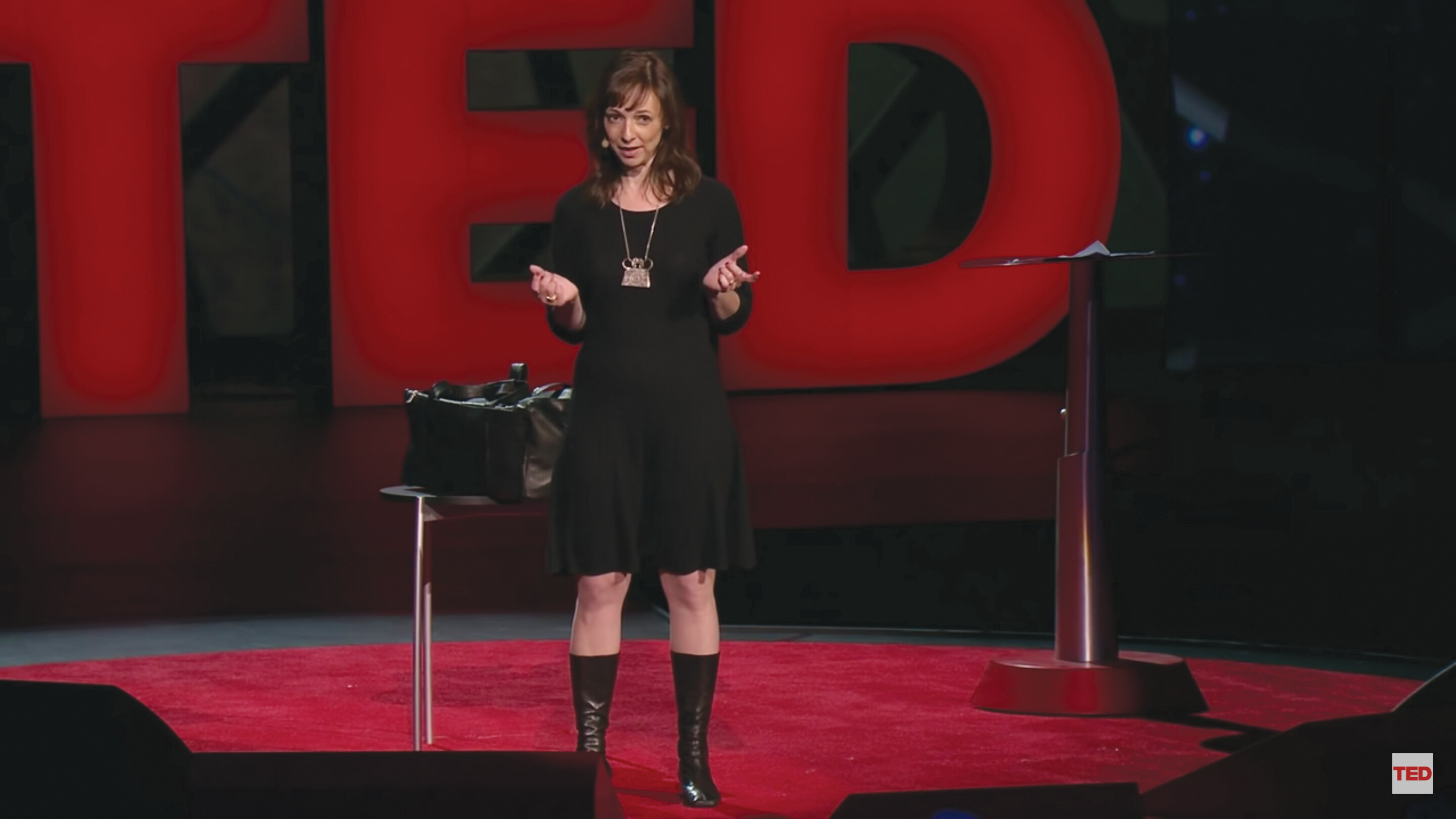 A screenshot of Susan Cain’s TED talk. She stands on a stage in an auditorium next to table.