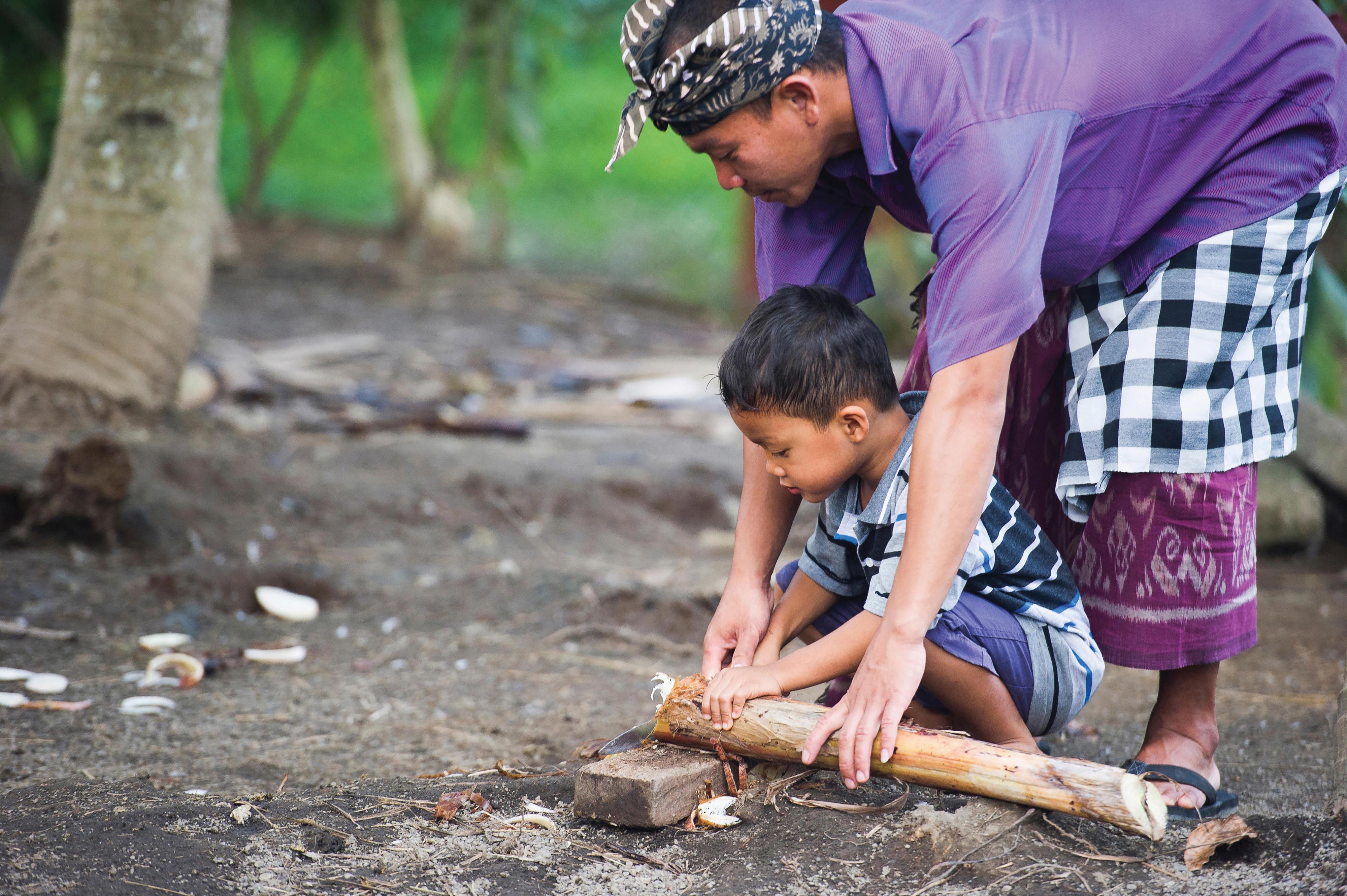 An Indonesian man teaches a young boy how to carve wood using traditional tools.