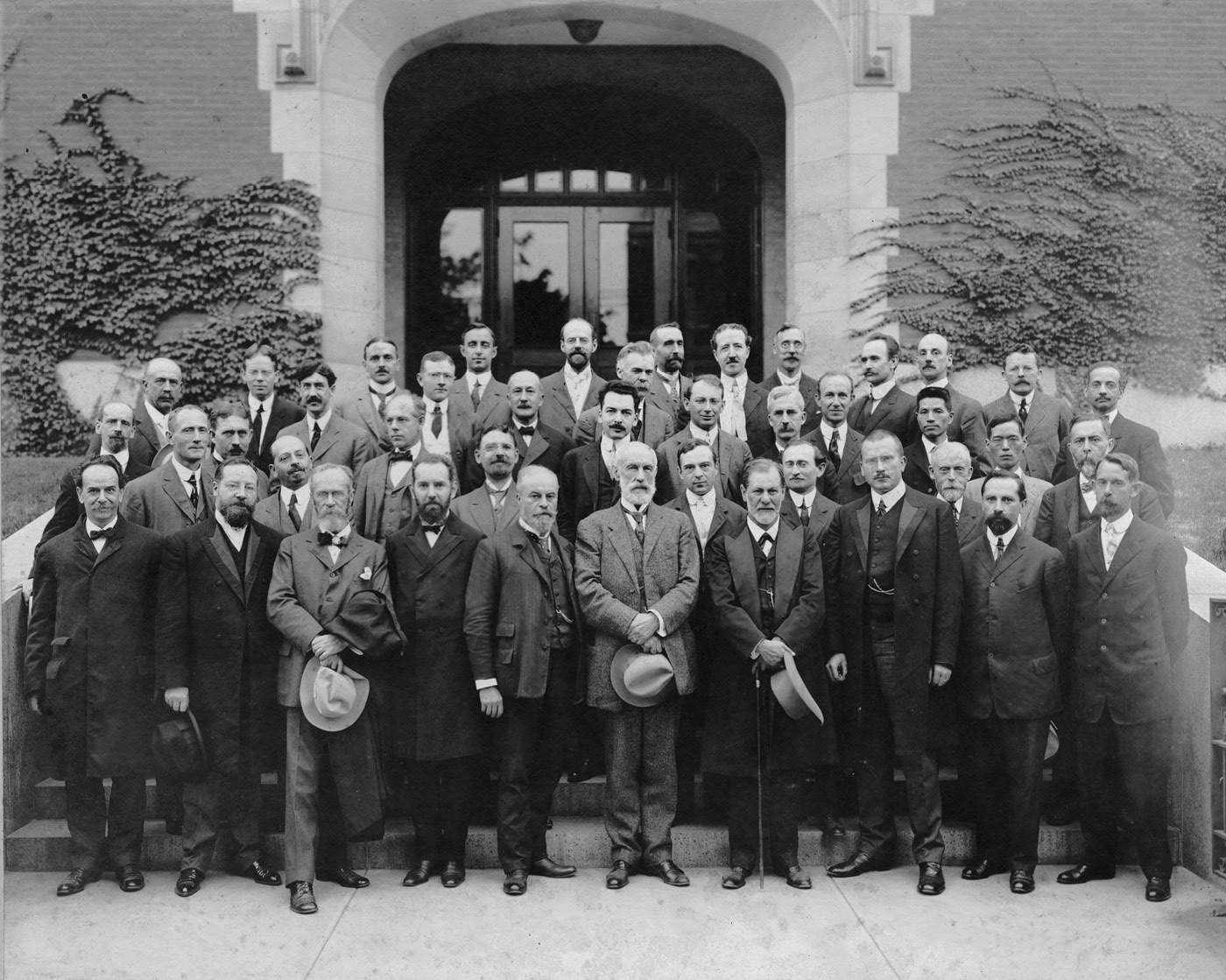 A large group of men wearing suits and coats pose for a picture outside in front of a building.
