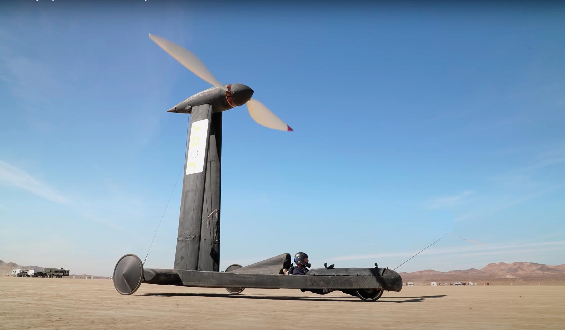 A full-size wind-powered vehicle, piloted by a helmeted figure, races across a flat desert floor.