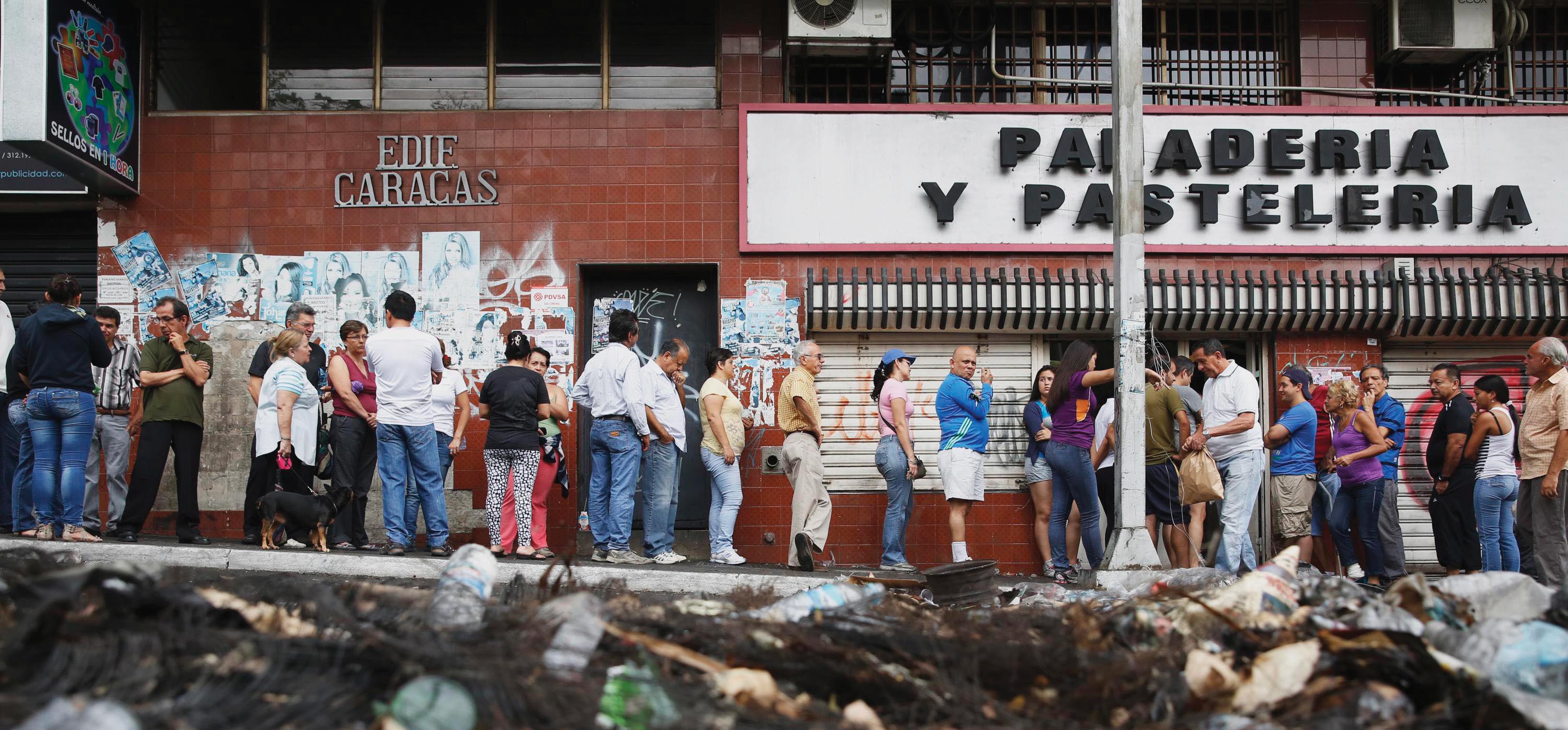 A line of people outside of a bakery with the sign: Panaderia y Pasteleria. There is a large pile of garbage in the street.