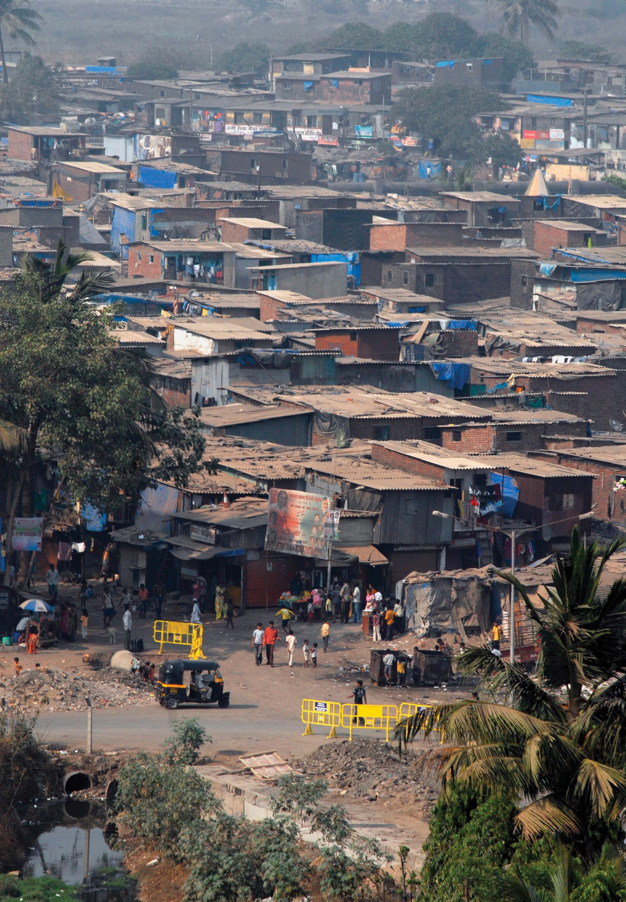 A winding dirt road forms the city street of a slum lined with clusters small shacks.
