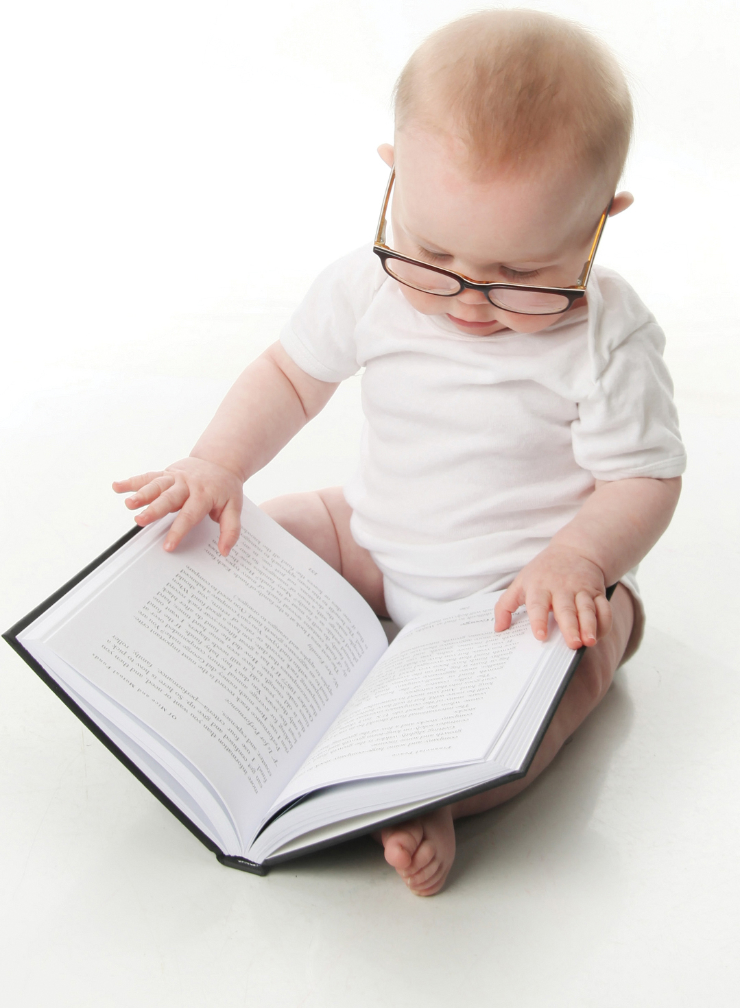 A baby wearing glasses and looking at an open book on its lap.