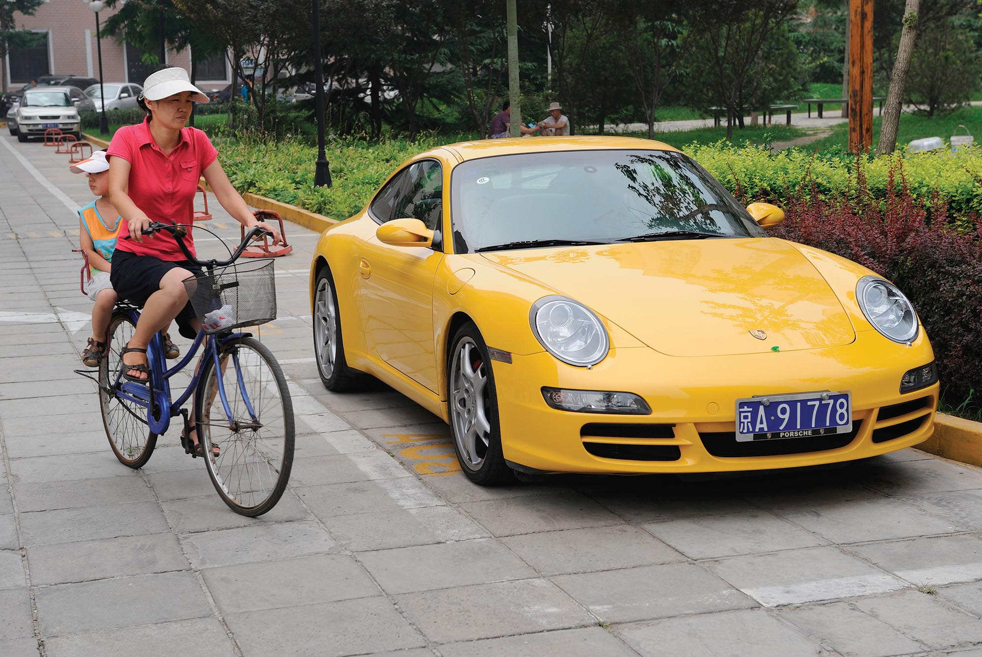 A yellow Porsche parked in a neighborhood. Next to the car is a mother riding a bicycle with a small boy sitting behind her.