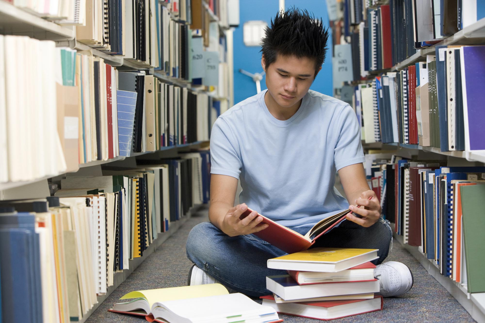 A student reading a book, while sitting in between shelves of books at a library.