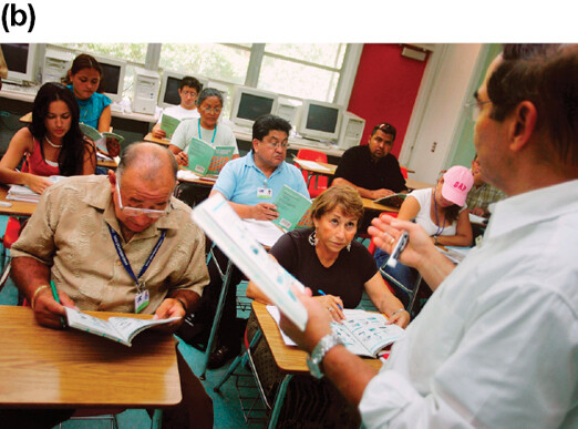 A man holding notes and lecturing a class of students, who are reading and taking notes.