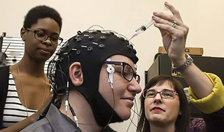 A man sits with a black helmet-like apparatus attached to his head. Electrodes are attached to the apparatus, and a woman prods an electrode using a syringe-like object.