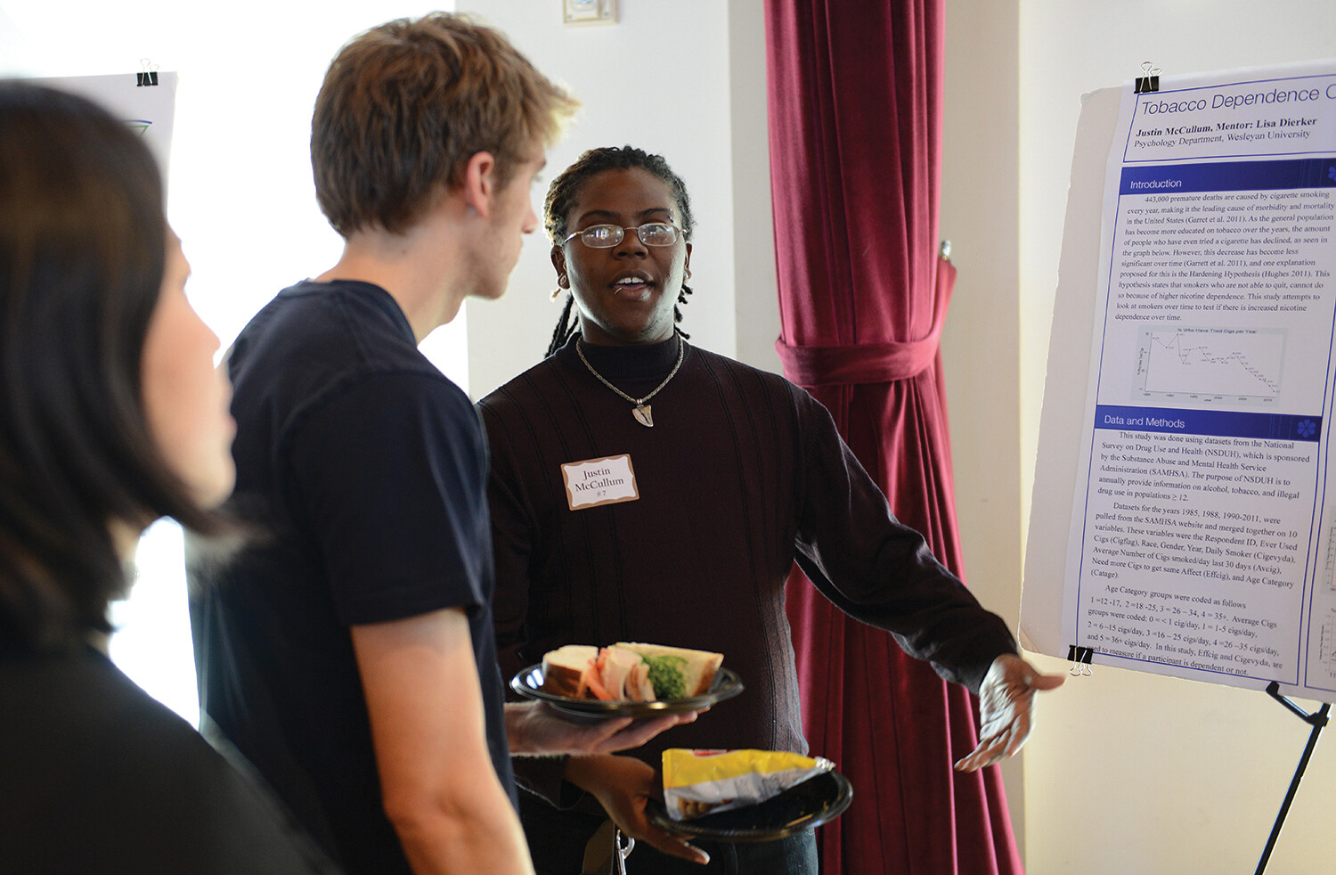 A woman stands beside a large poster on a board, and speaks to a man carrying a plate of food.
