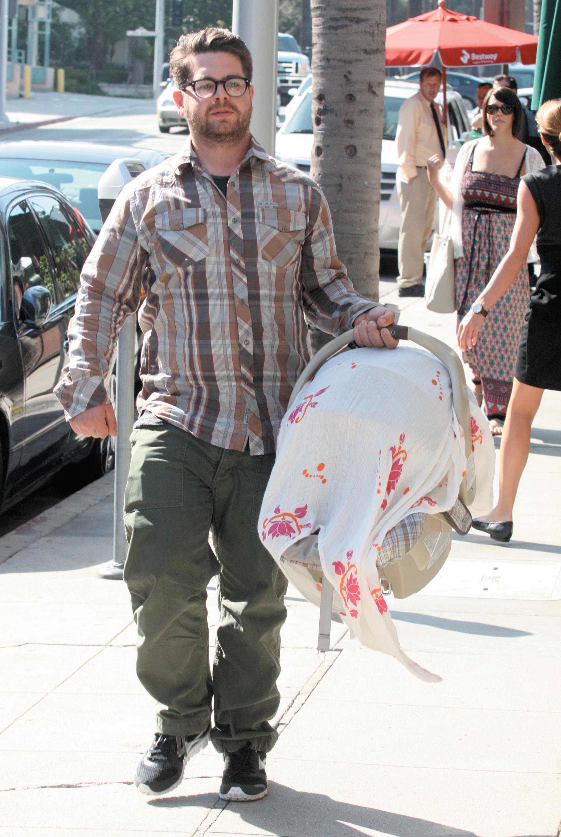 A photo of a man walking down the street with a baby carrier in his hand.