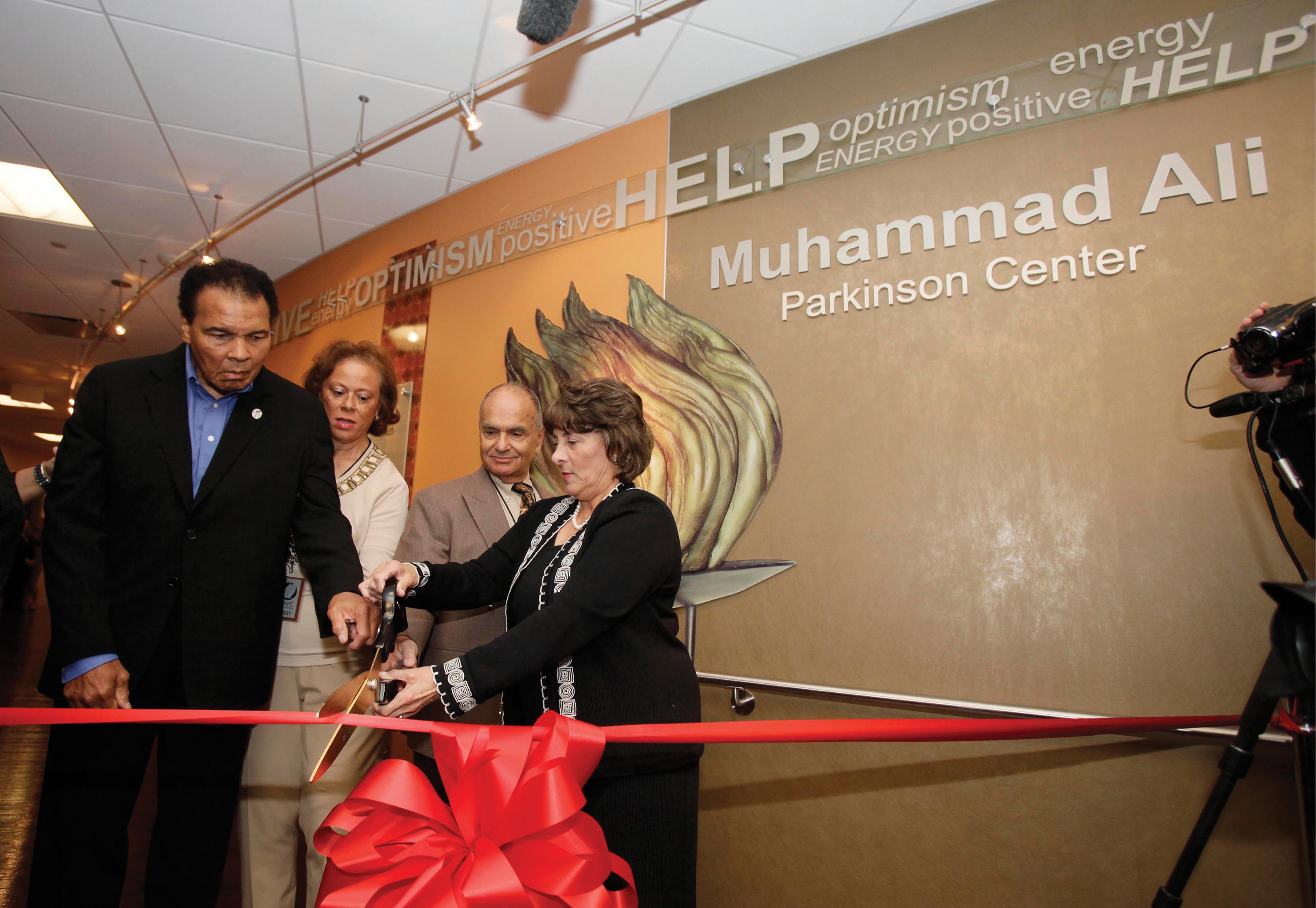 A photo depicting Muhammad Ali cutting a large red ribbon in the dedication of a Parkinson’s center named for him.