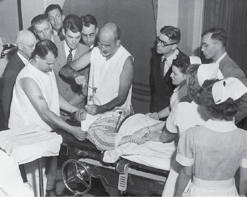 A black and white photograph of a man giving a surgical demonstration to 8 men in suits and three nurses or orderlies.