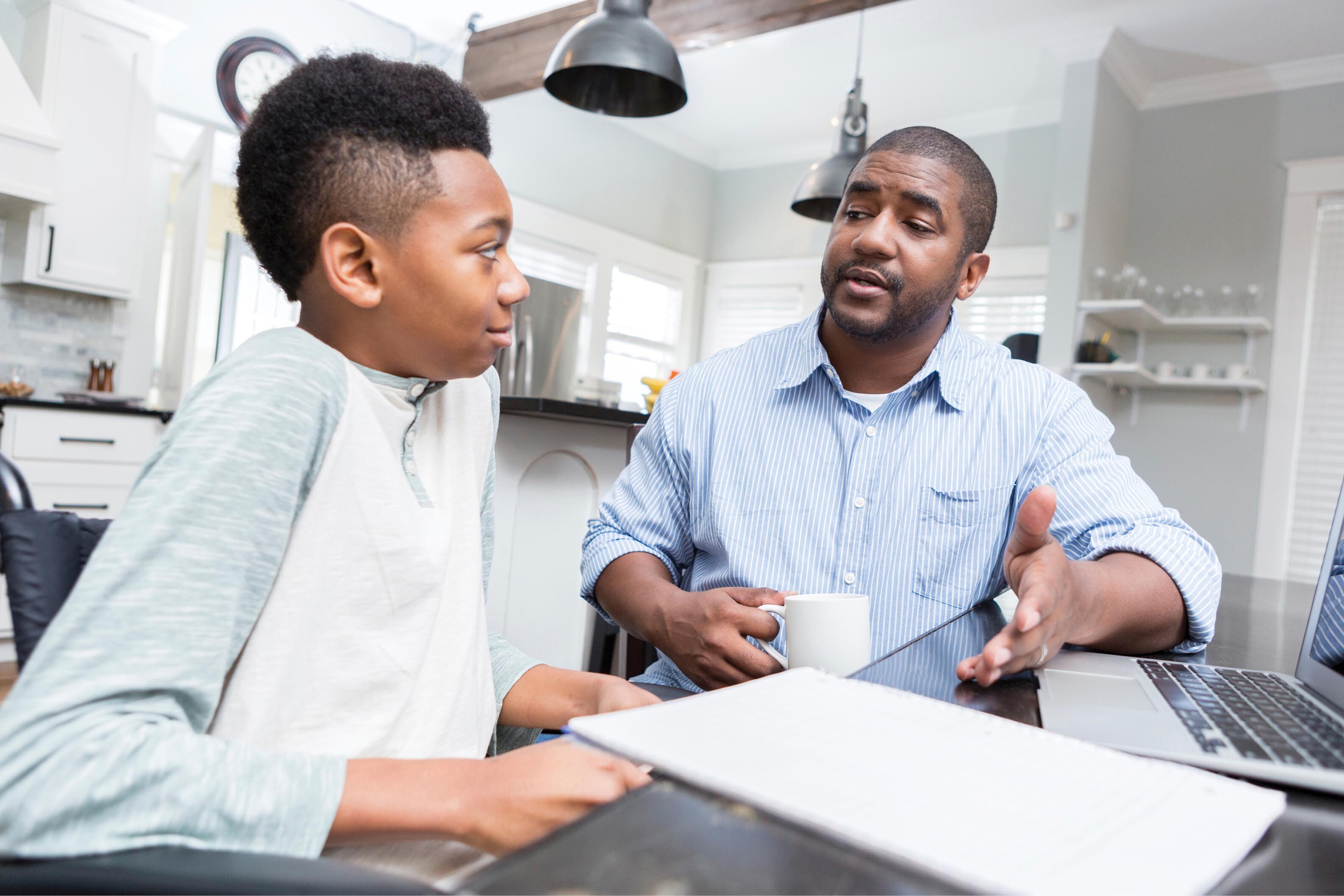 A photo shows an interaction between a boy and his father. They both sit at a table. A book and a laptop are present on the table.