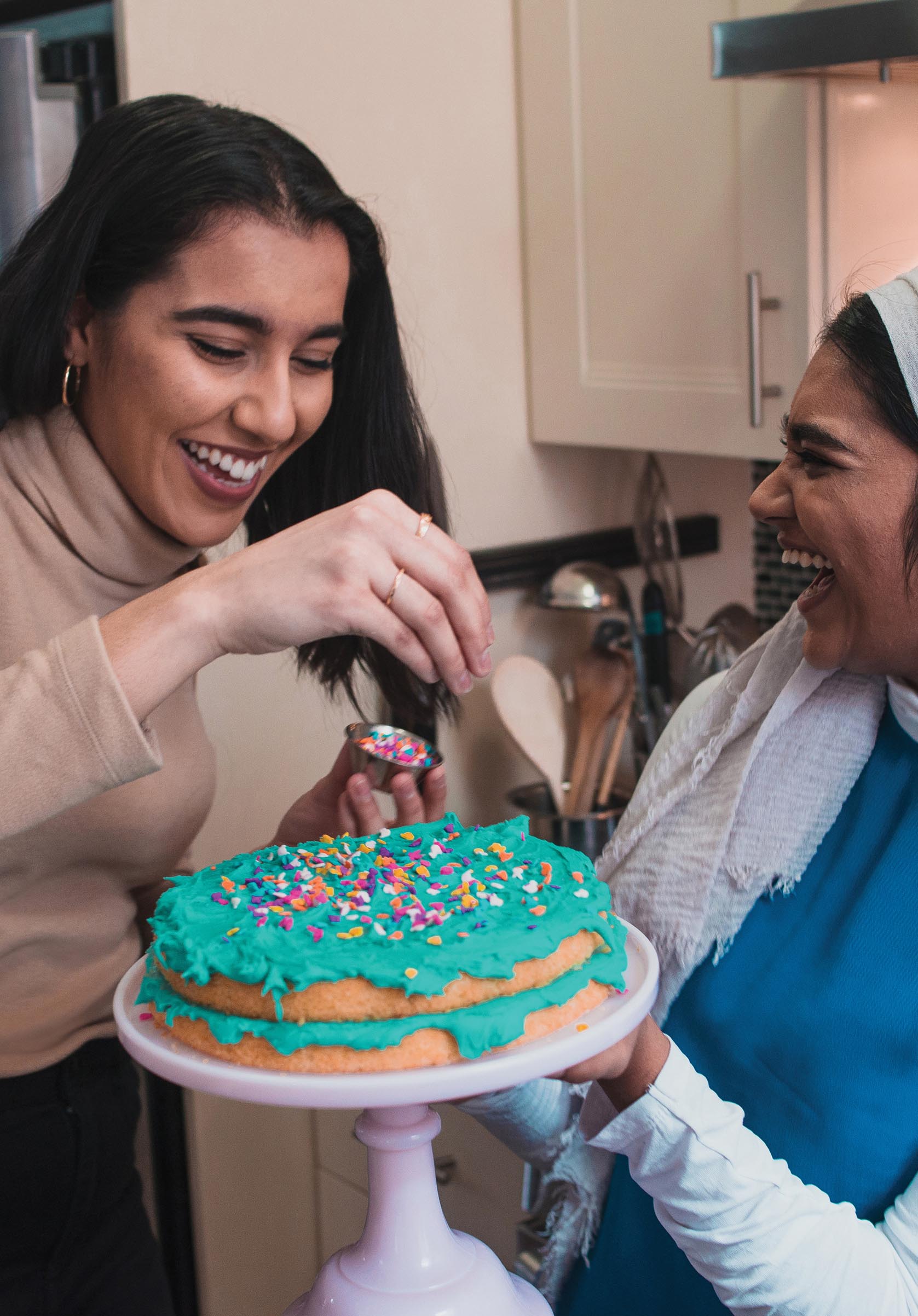 Two laughing women stand beside each other. One woman holds a cake, and the other woman decorates the cake with sprinkles.