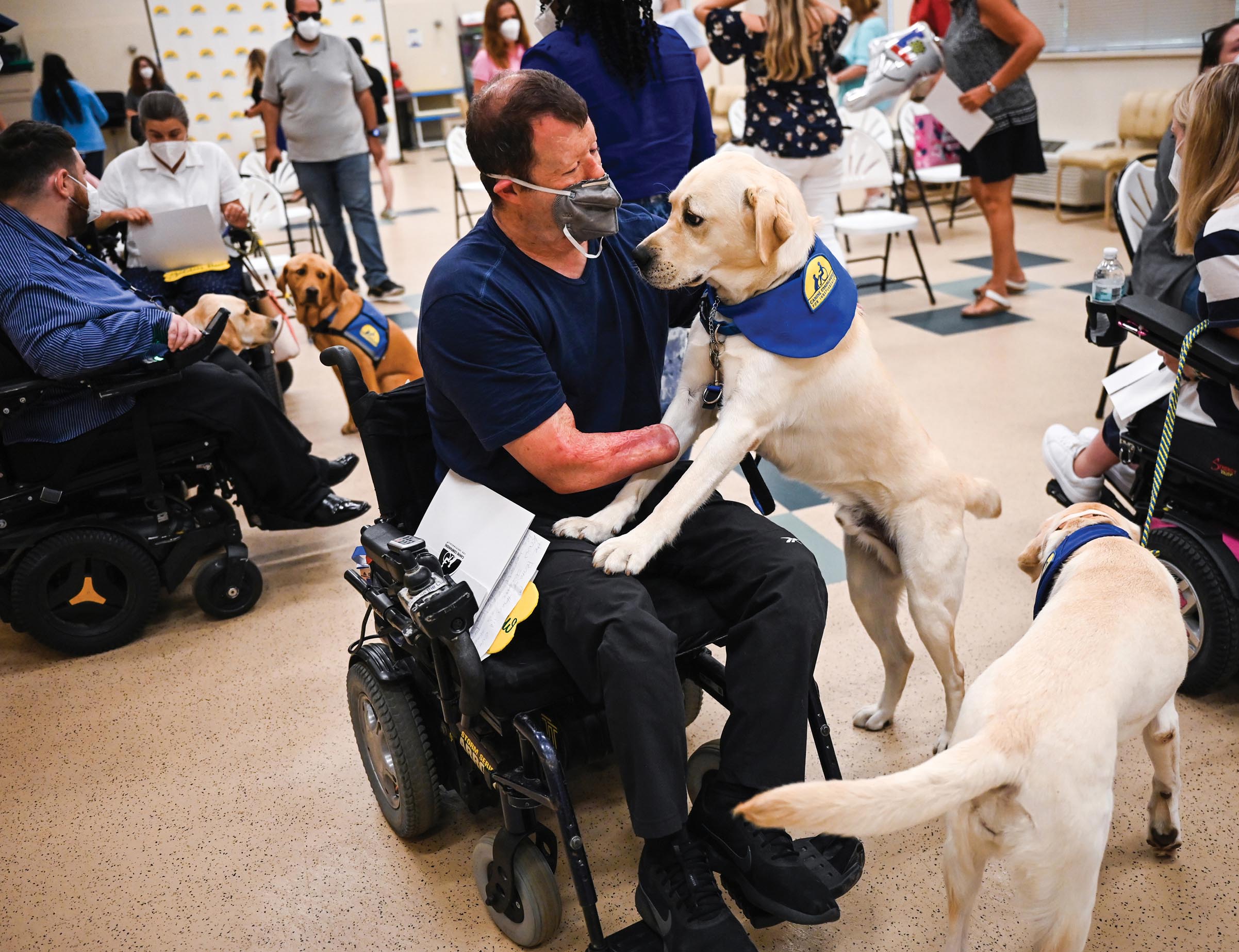 A man on a wheelchair, wearing a mask, pets a dog that leaps onto his lap. He is surrounded by other people on wheelchairs and dogs.