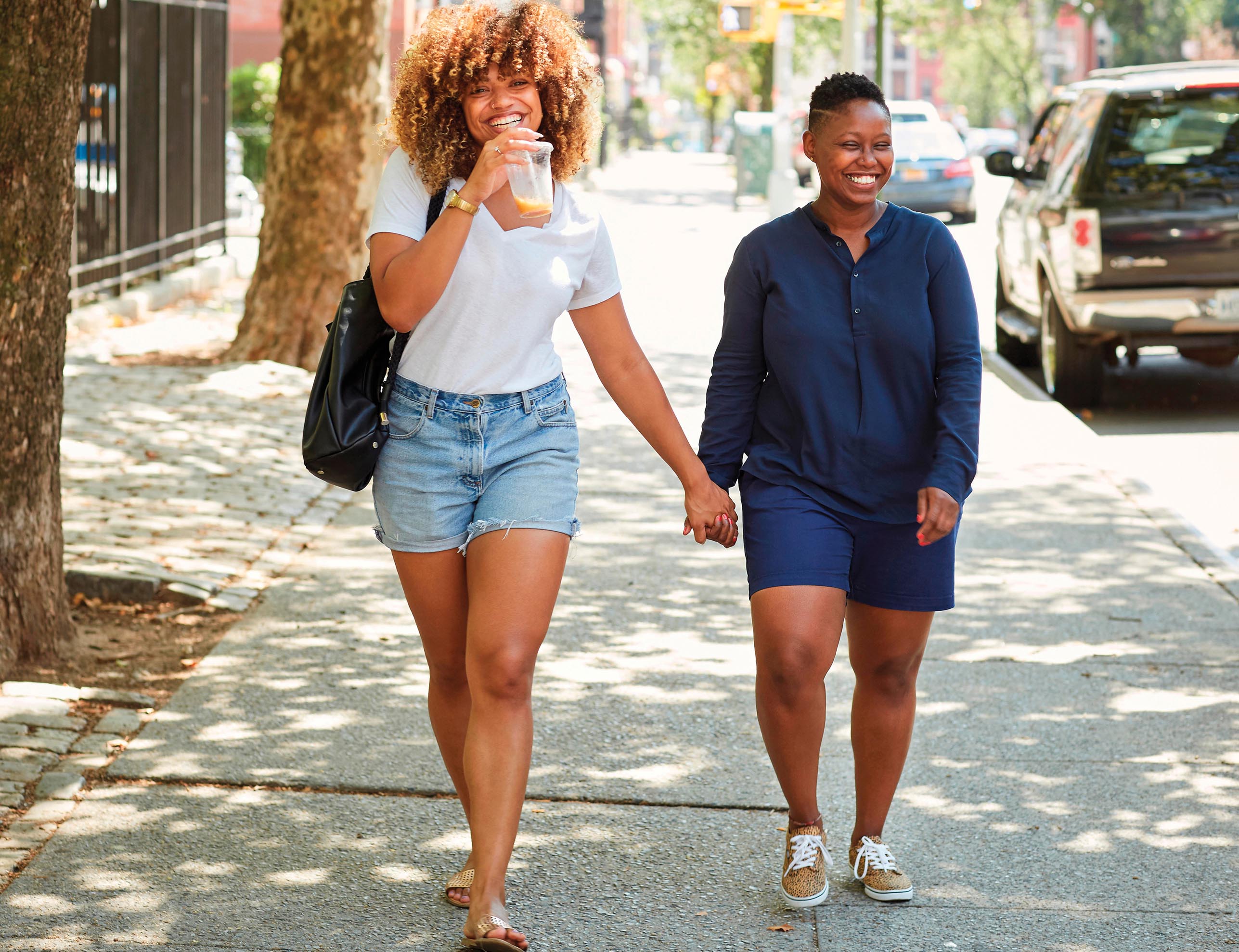 A photo of two women holding each other’s hands and walking on the pavement at the sides of a road.