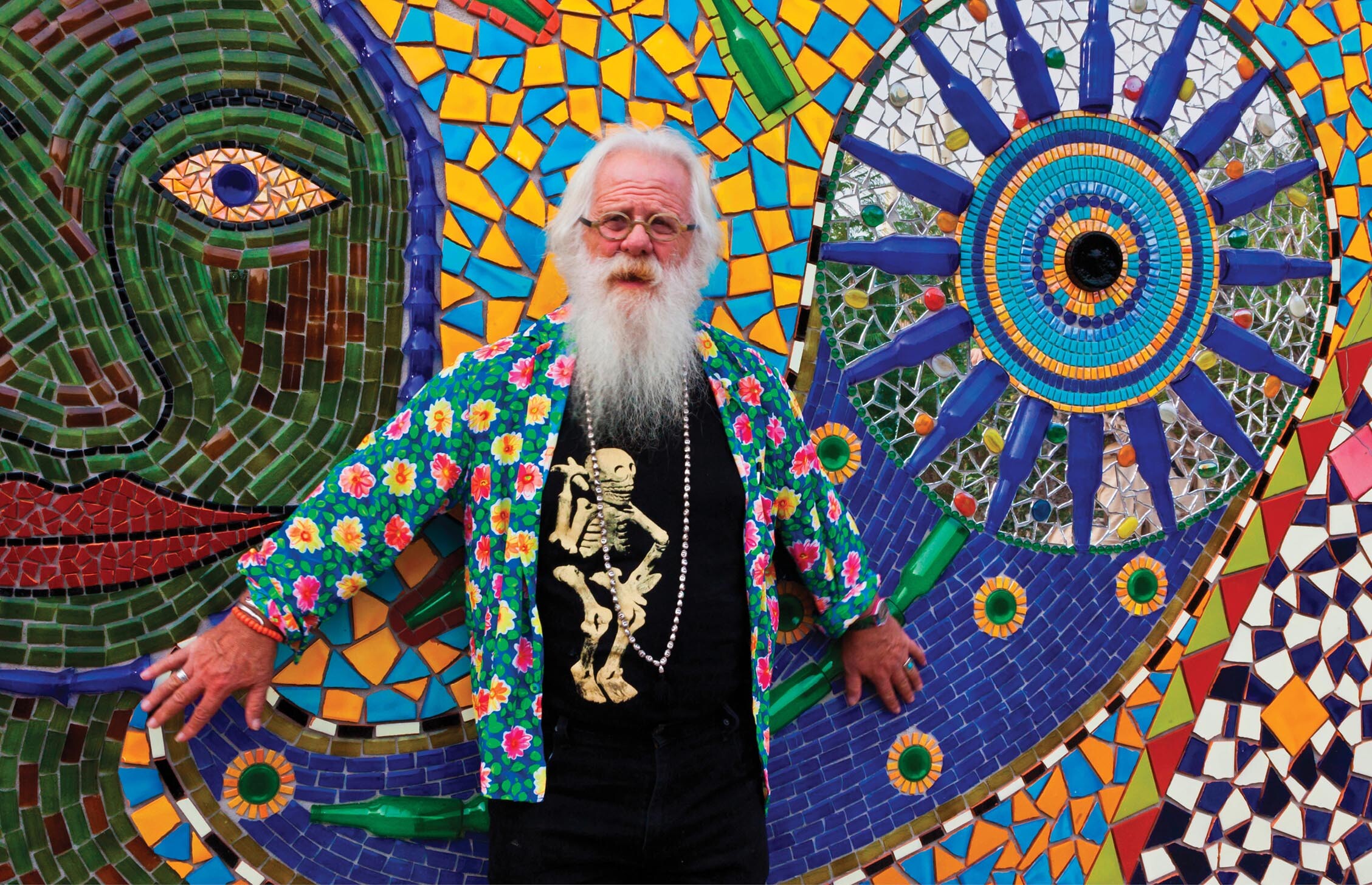A photo of an old man with a long beard and a long beaded chain around his neck leans against a wall decorated with many artworks.