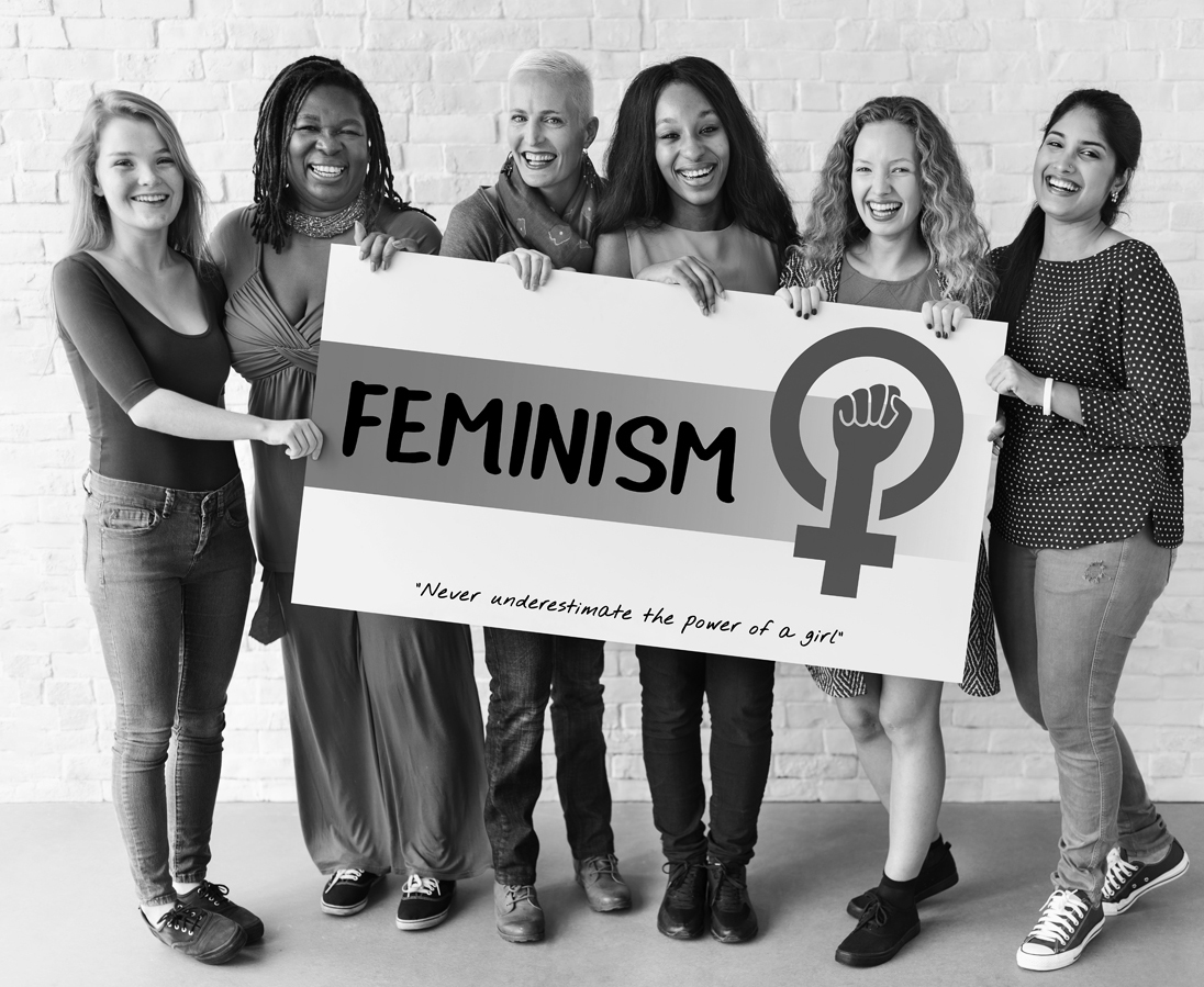 A photo shows a group of women, belonging to different ethnicities, smiling and holding a board that reads “Feminism.”