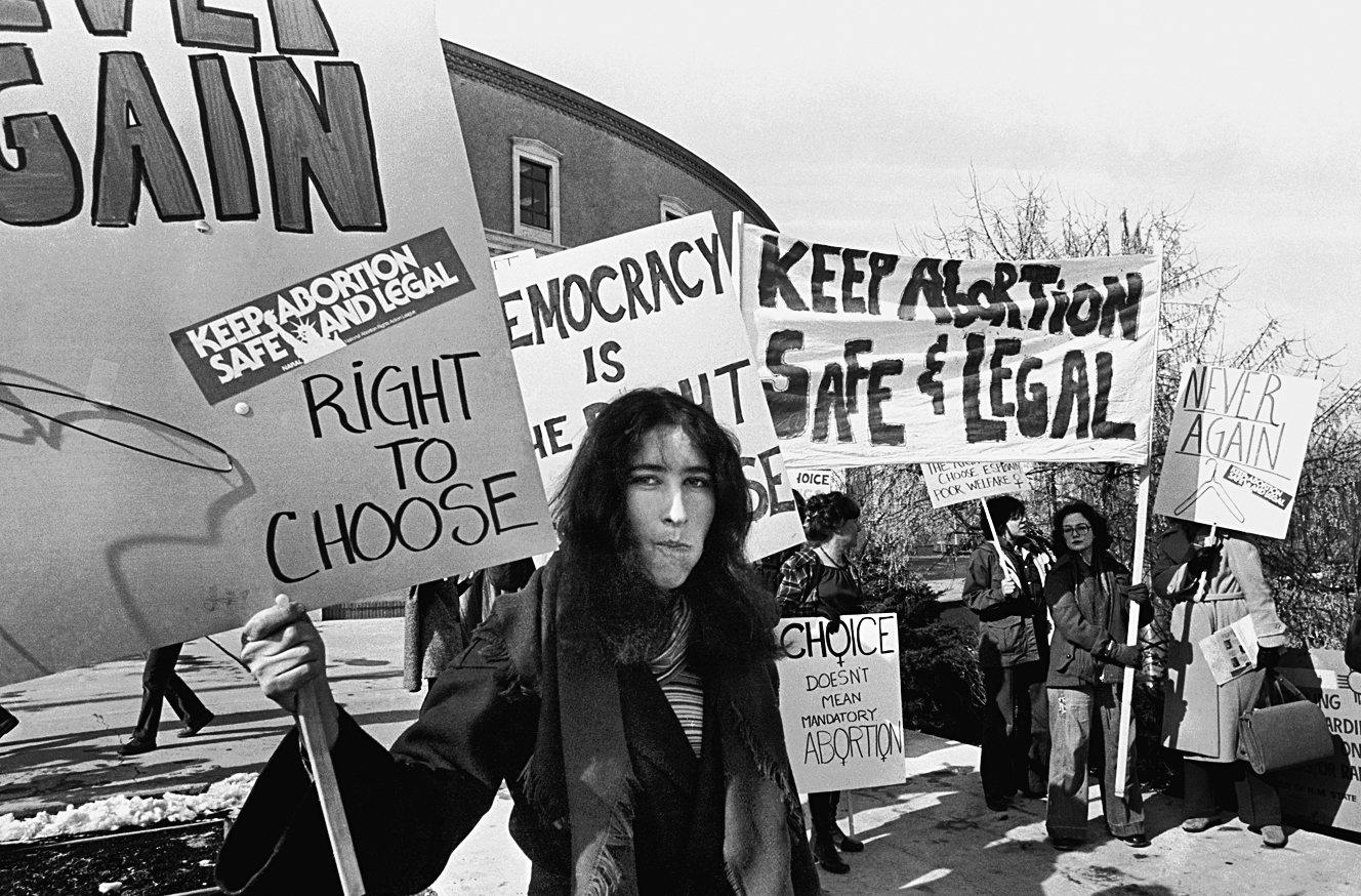 A photo shows a group of women protesting in support of legal abortion. They hold banners and placards that read “Keep abortion safe and legal,” “Choice doesn’t mean mandatory abortion,” and “Never Again.”
