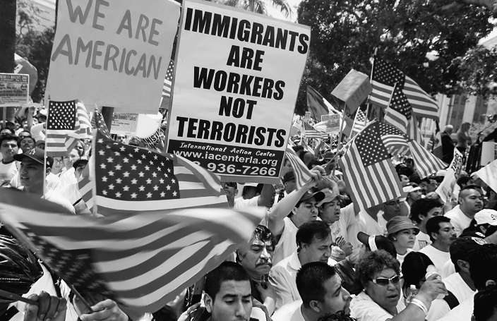 A photo shows a crowd of protesters waving American flags and holding signs in favor of immigration. Signs read “We are American” and “Immigrants are workers not terrorists, International Action Center.”