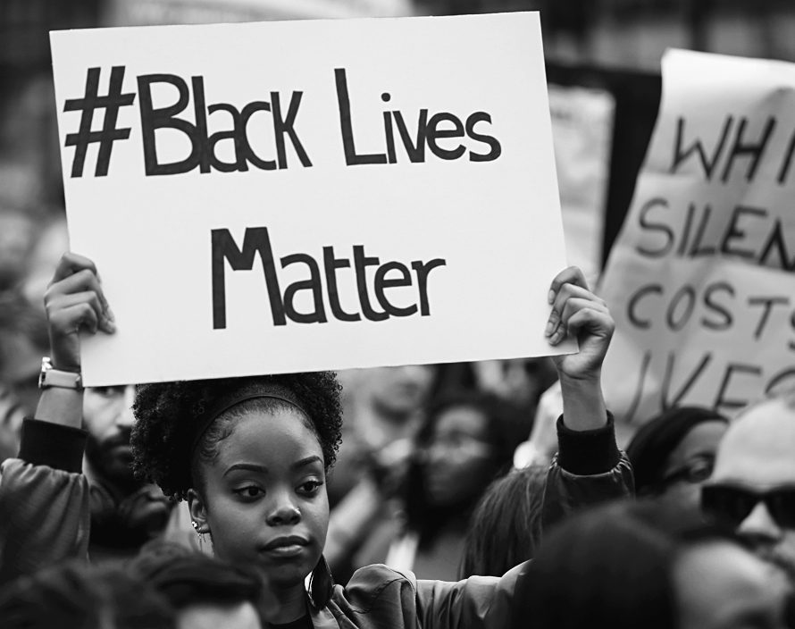 A photo shows an African-American woman standing among a crowd and holding a poster that reads “hashtag Black lives matter.”