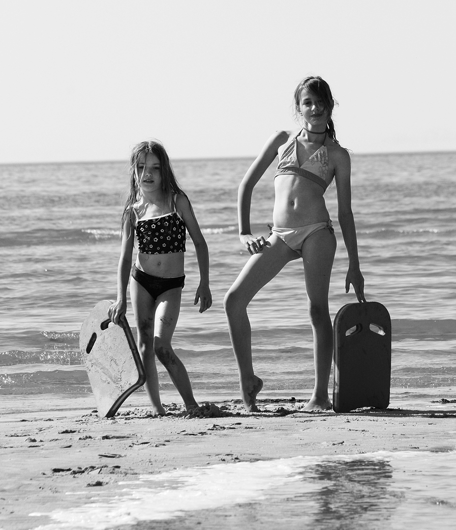 A photo shows two young girls at the beach dressed in swimsuits, each holding a kickboard.