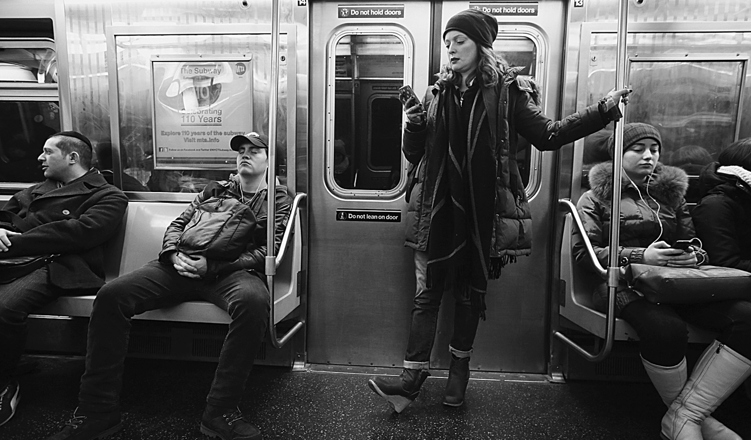 A photo shows a man seated in a subway car with his legs wide apart, taking up two seats. A woman stands next to him.