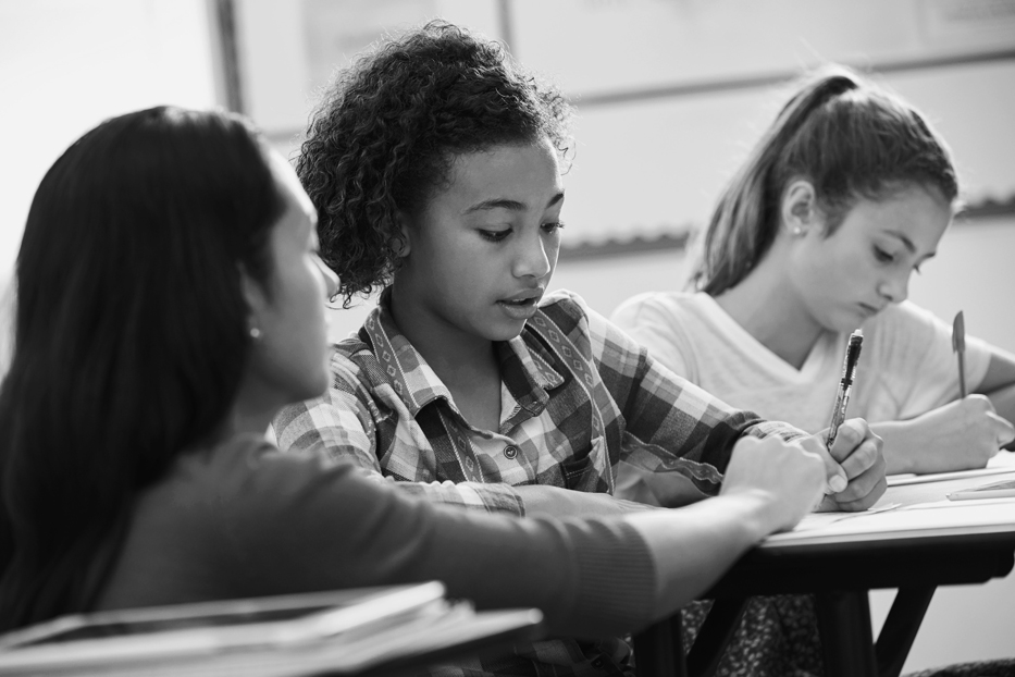 A photo shows a female teacher helping two teenage girls with their studies.