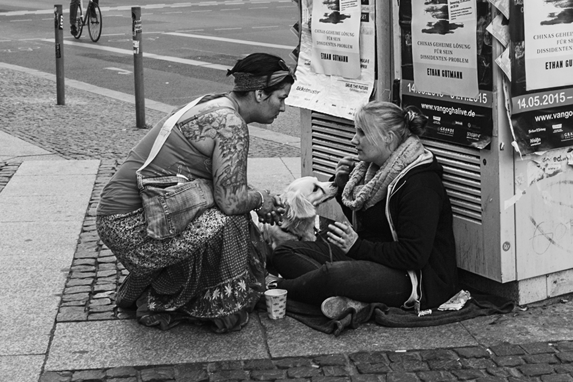 A photo shows a woman listening to a needy woman seated on a matt on the sidewalk of a city street.