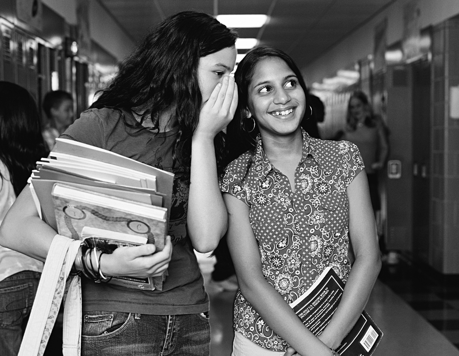 A photo shows a teen girl cupping her hand and whispering into the ear of a girl next to her in a school hallway.