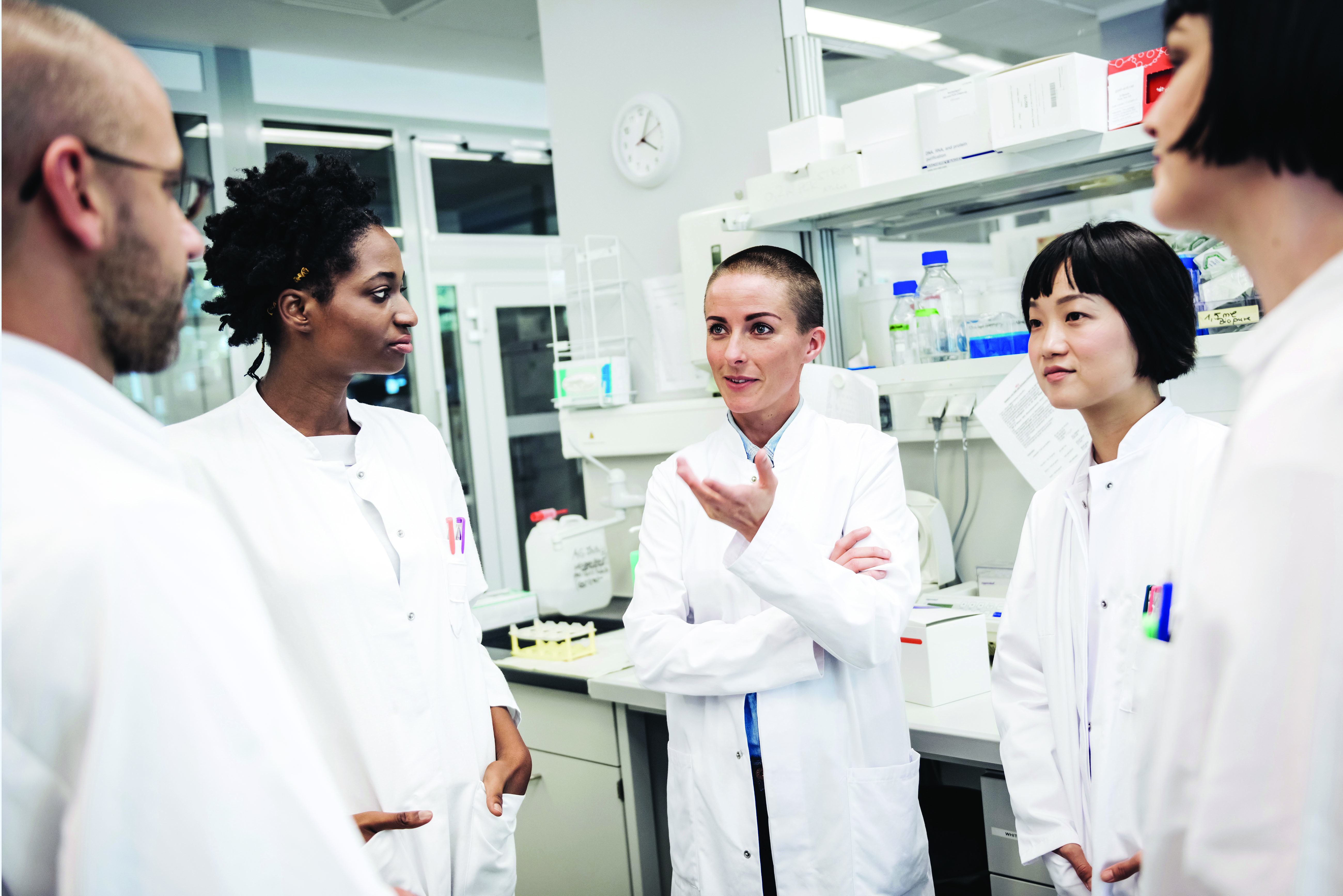 A photo shows five scientists standing dressed in white lab coats engaged in a conversation in a lab.