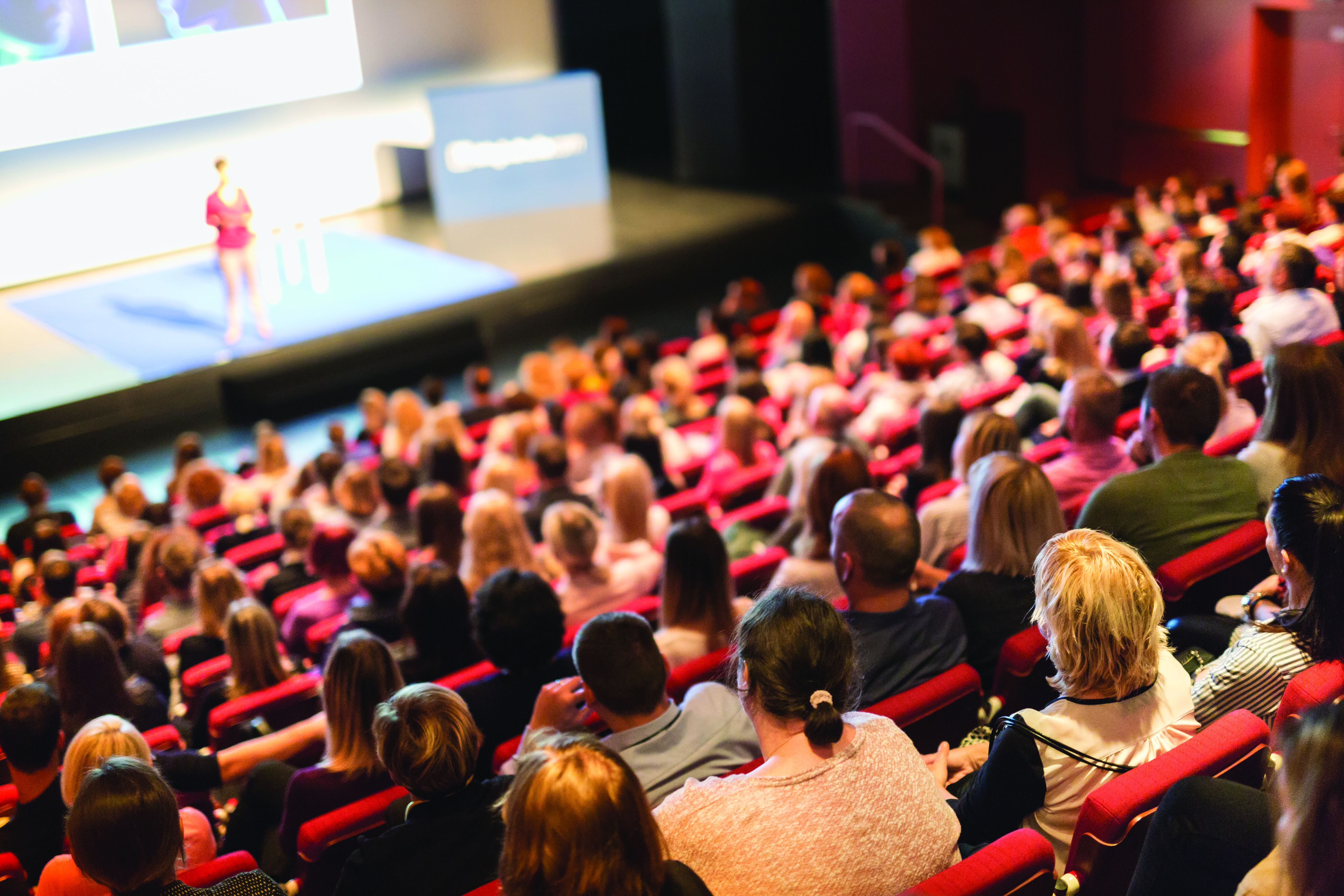 A photo shows an auditorium full of people with a person standing on stage.