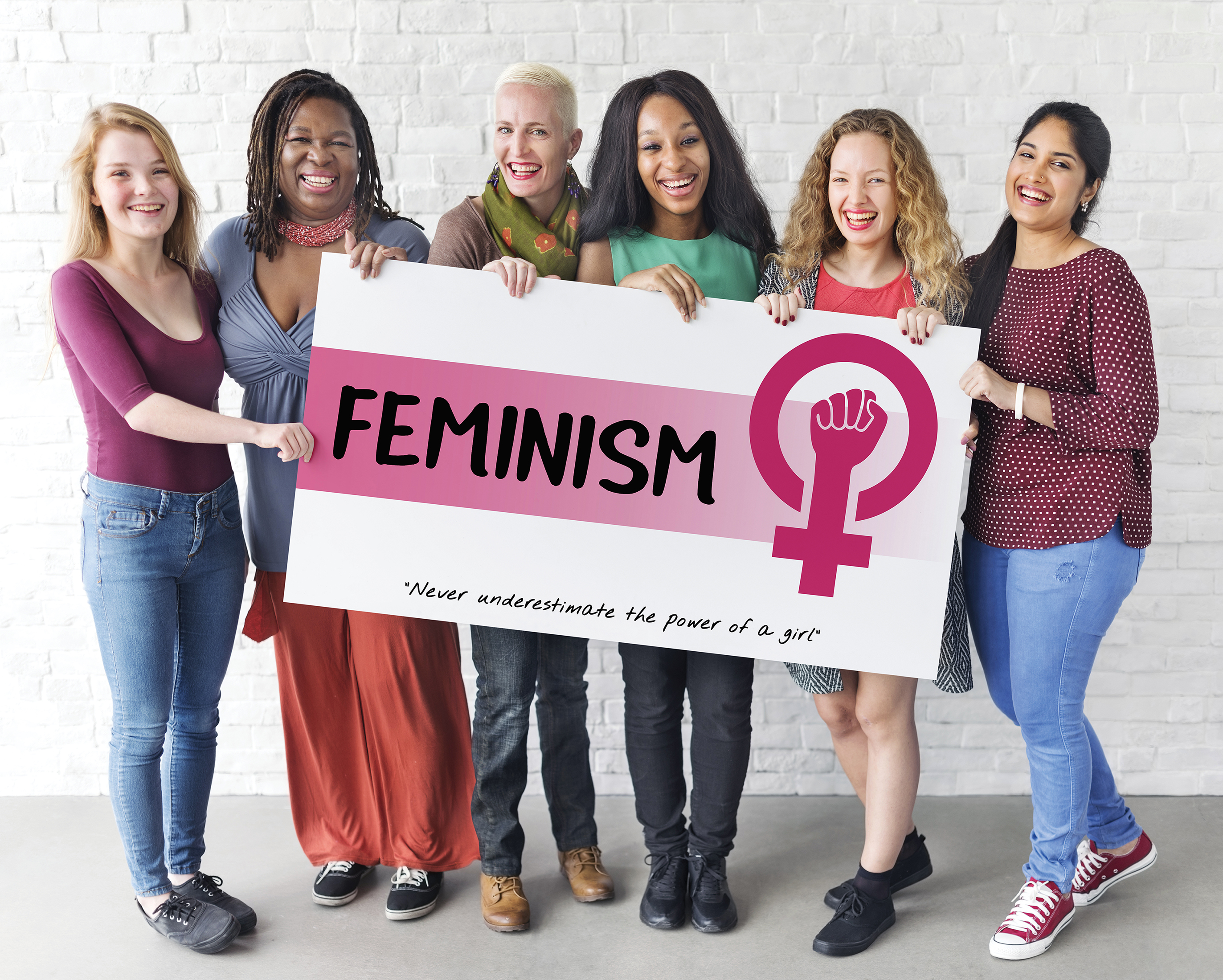 A photo shows a group of women, belonging to different ethnicities, smiling and holding a board that reads “Feminism.”