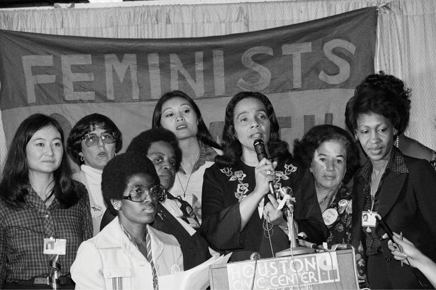 A black and white photo shows a woman speaking at a podium with a group of female activists standing with her.