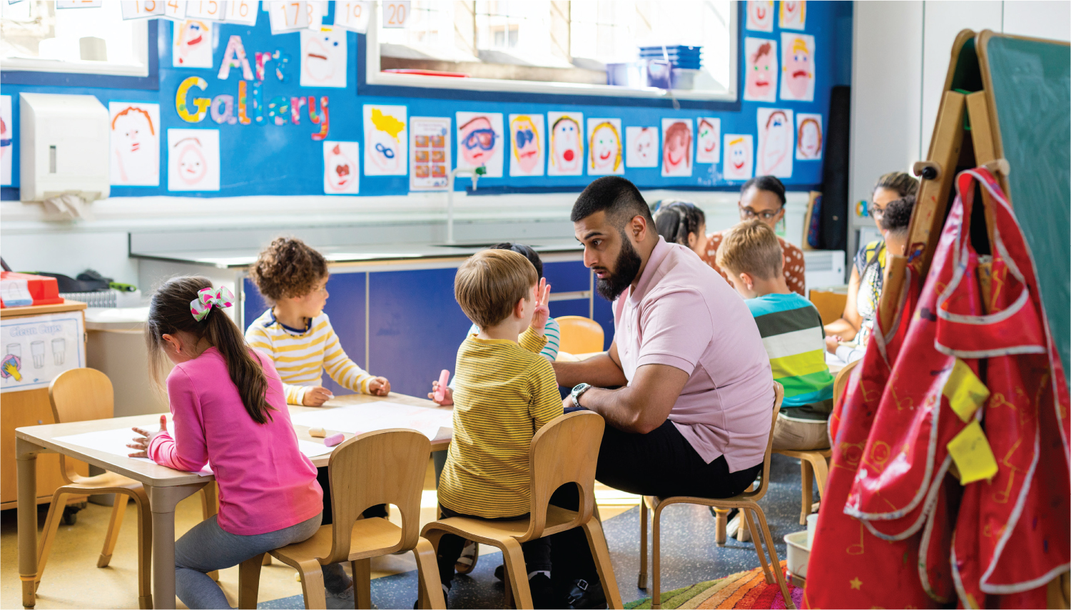 A photo shows a male preschool teacher in a classroom interacting with a child at a kid’s table with other children writing.