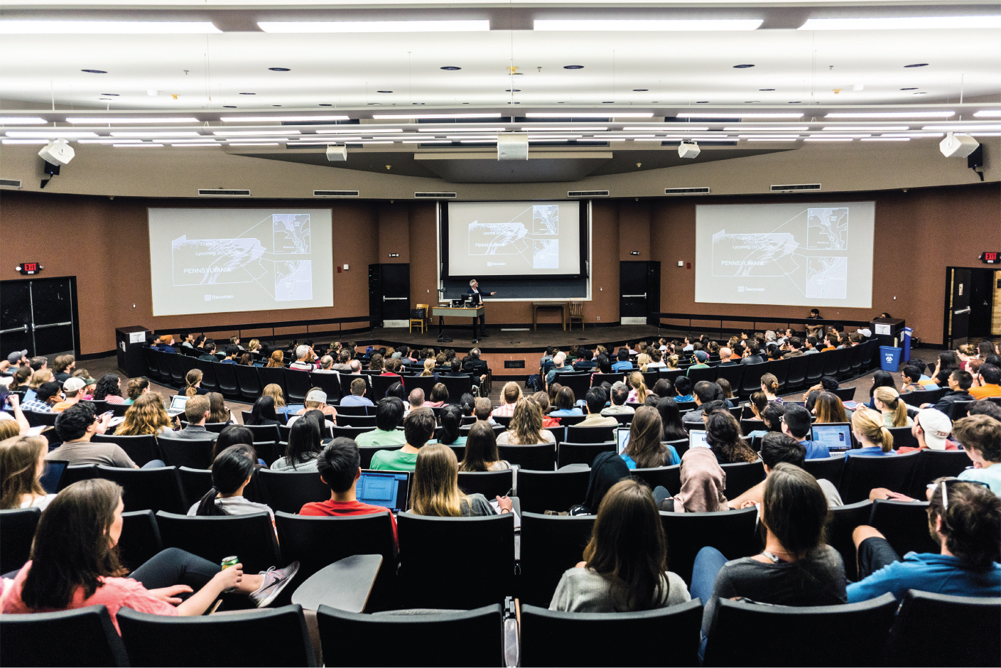 A photo shows a college classroom full of students, a professor giving a lecture, and three screens with lecture slides.