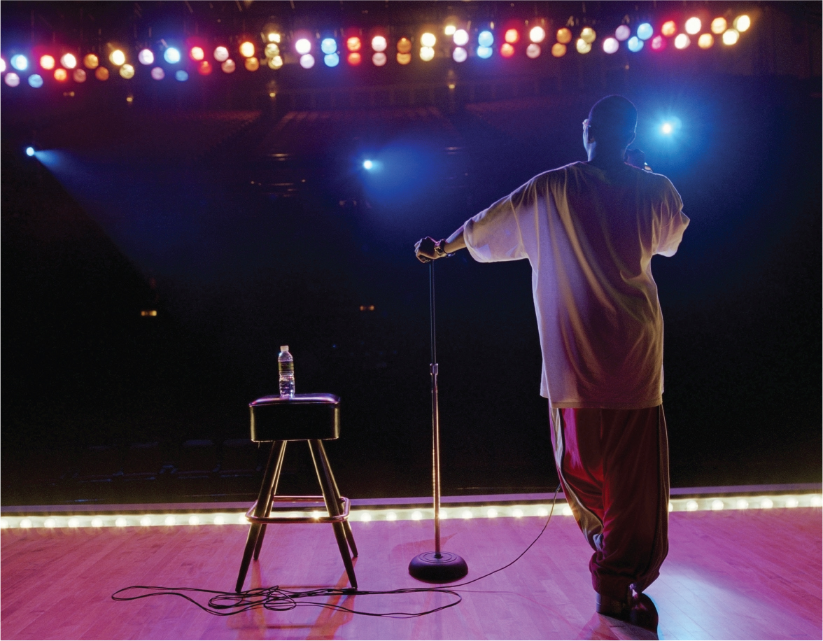 A photo shows the back view of a comedian on a stage with bright lights, holding a microphone speaking to a darkly lit room.