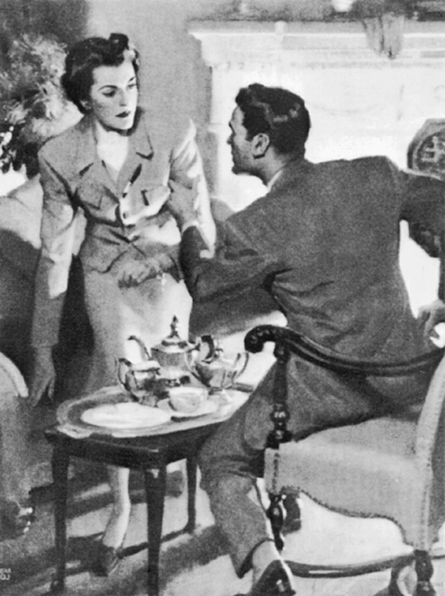 A black and white picture shows a man and woman at a table set with tea in front of a fireplace.