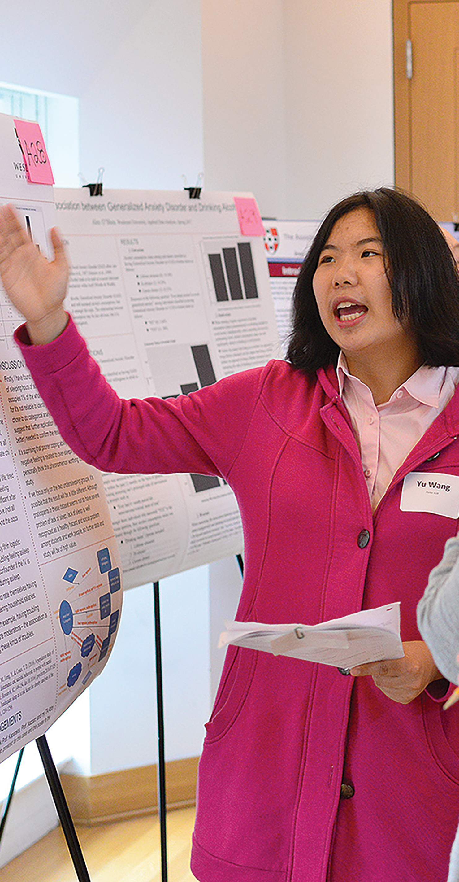 A researcher is shown talking while holding papers in her hand and gesturing at a presentation board.