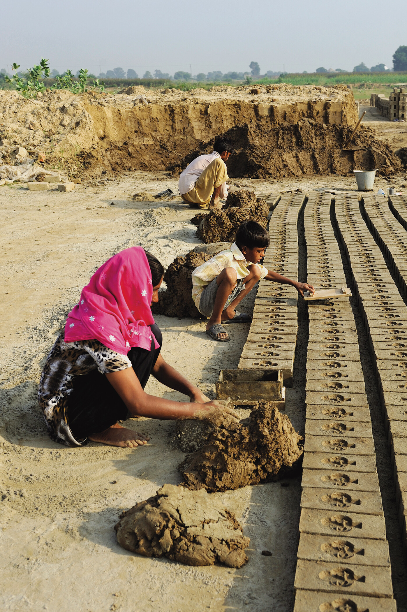 Two young boys and a young girl crouch down to mold bricks.