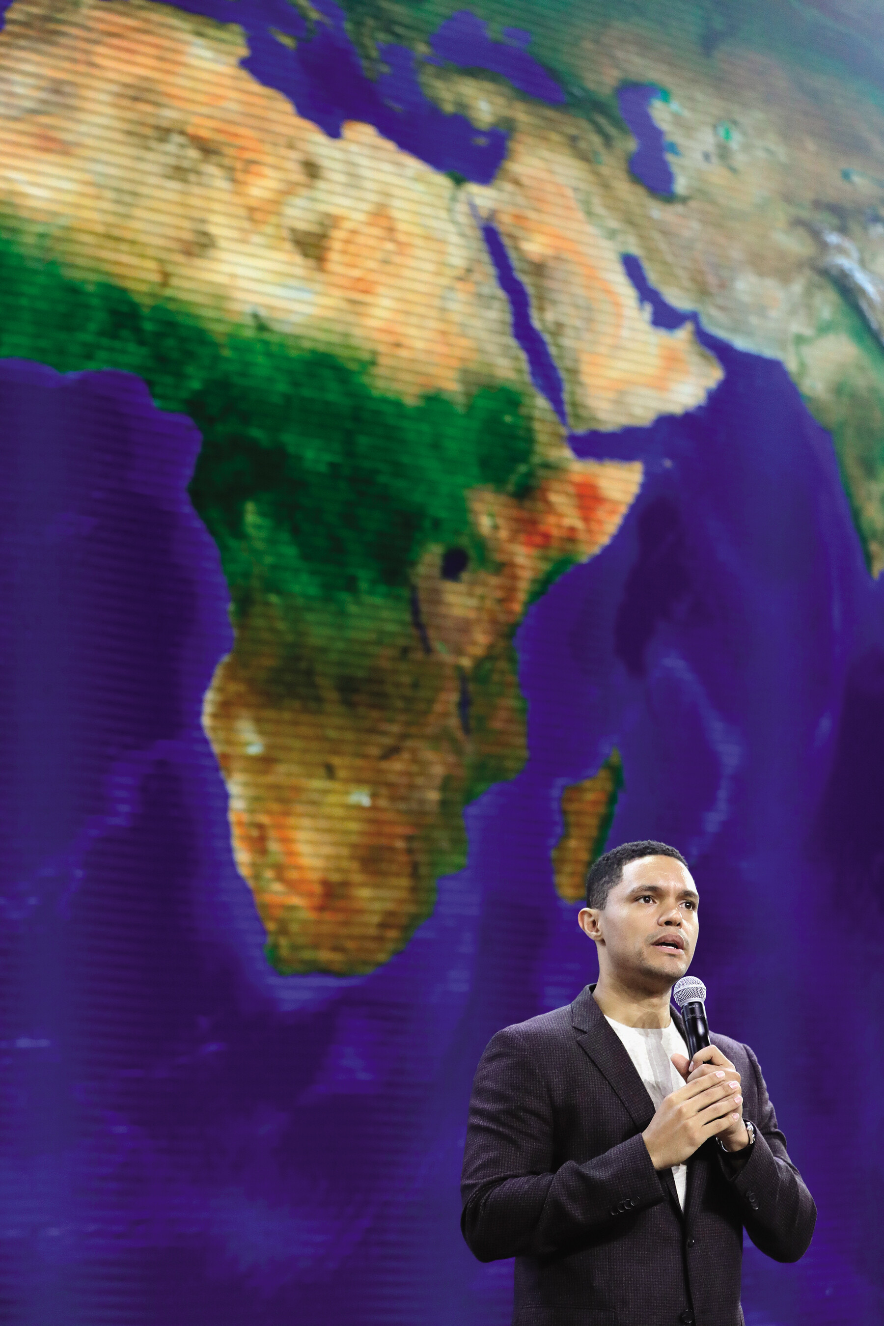 Trevor Noah holds a microphone and stands in front of a screen.