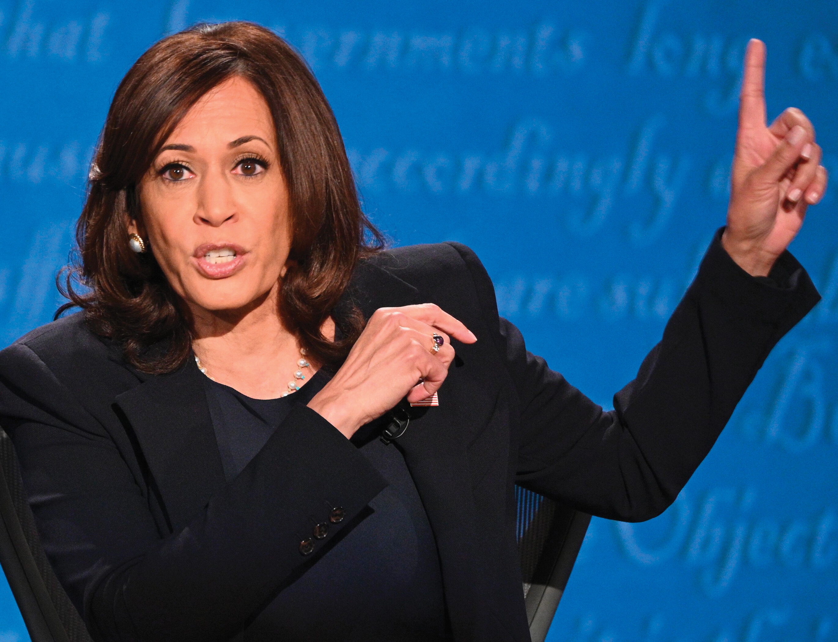 A woman sits onstage looking straight forward. She is holding up her left index finger to someone off-camera next to her.