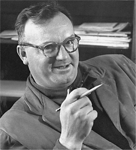 A black-and-white photo of a white man wearing glasses leaning back in his chair, pencil in hand.