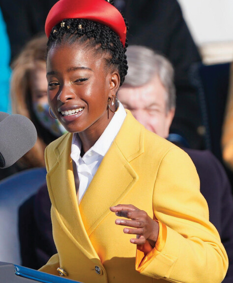 Amanda Gorman speaks at a podium, gesticulating with her hands. She wears a bright yellow coat and a red satin headband.