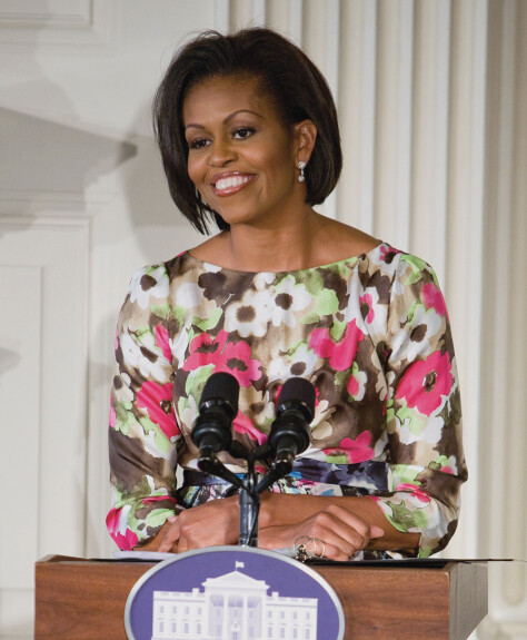 Michelle Obama smiles while standing at a podium with a placard of the White House attached to it.