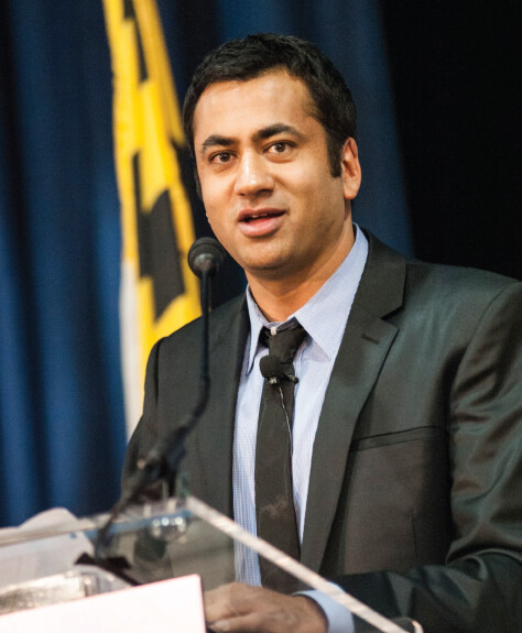 Kal Penn speaks at a podium with a black-and-yellow Maryland flag behind him.