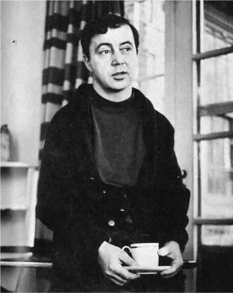 A black-and-white portait of a white man with dark hair leaning on a desk behind him and holding a coffee cup and saucer.